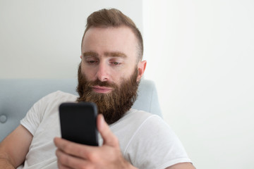 Pensive bearded phone user focused on phone screen. Young man in casual resting on couch and using smartphone. Phone using concept