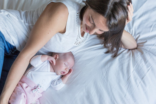 Mother And Baby At Home Lying On Bed And Relaxing. Baby Girl Sleeping And Mother Looking At Her. Motherhood Concept