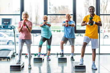 happy senior multicultural sportspeople synchronous exercising with dumbbells on step platforms at...