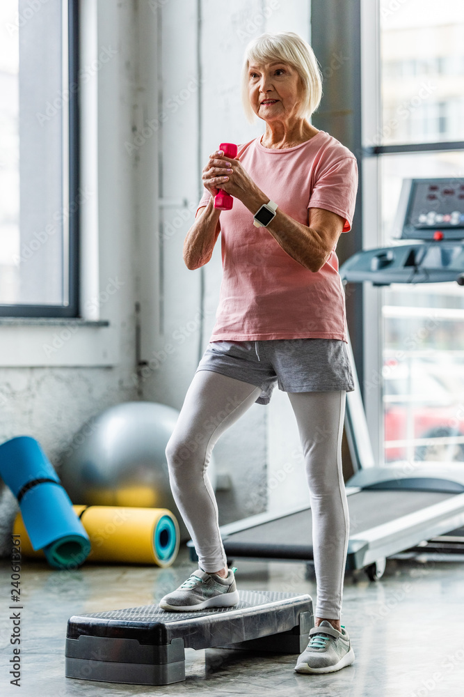 Wall mural senior sportswoman with smartwatch making exercise with dumbbell on step platform at gym