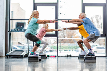 side view of mature athletes synchronous doing squats on step platforms at gym