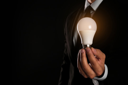 Businessman holding lamp bulb against dark background, closeup. Space for text