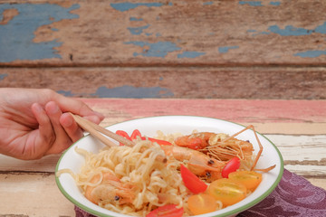 Female hand using wooden  chopsticks to eat instant  noodles with shrimp, tomato cherry and  chili on wooden background