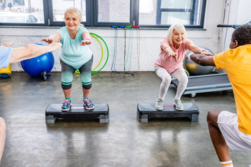 smiling senior multiethnic sportspeople synchronous doing squats on step platforms at gym