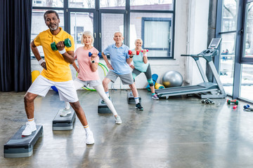 multiethnic senior sportspeople synchronous exercising with dumbbells on step platforms at gym