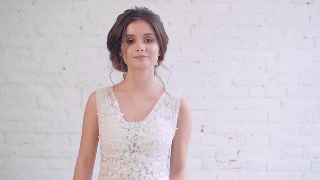 attractive image of dark-haired bride with neat, gentle hairstyle, goes and looks directly at camera, spinning in long white elegant dress, portrait shooting of girl near white-washed brick wall