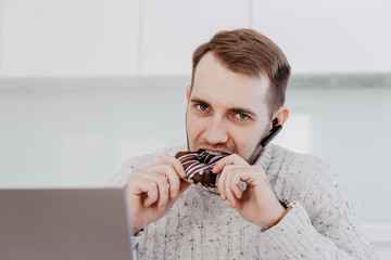 A man with a beard is sitting at the table in the white kitchen and eating a chocolate donut for lunch