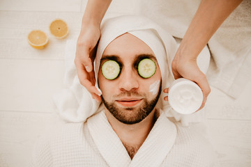 Relaxed bearded young man in a towel on his head being in spa salon with moisturizer cucumber and...