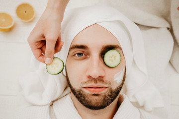 Relaxed bearded young man in a towel on his head being in spa salon with moisturizer cucumber and mask on the face