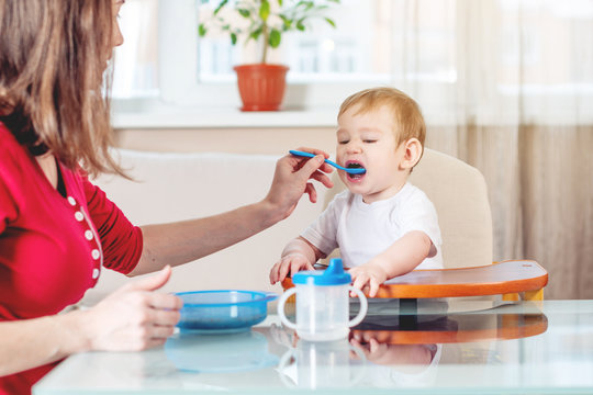 Mom Feeding The Baby Holding Out Her Hand With A Spoon Of Food In The Kitchen. Emotions Of A Child While Eating