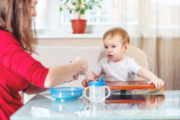 Mother feeding the baby holding out her hand with a spoon of porridge in the kitchen. Emotions of a child while eating