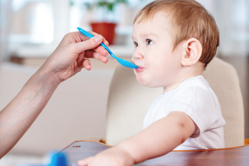 Mom feeding the baby holding hand with a spoon of porridge in the kitchen. Emotions of a child while eating