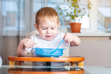 Little baby boy learning to eat with a spoon himself at the kids table in the kitchen. Concept of healthy baby food