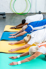 Fototapeta premium side view of multicultural senior athletes synchronous exercising on fitness mats at gym