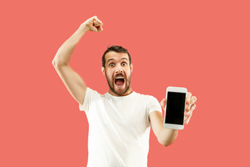 Young handsome man showing smartphone screen over coral background with a surprise face. Human...