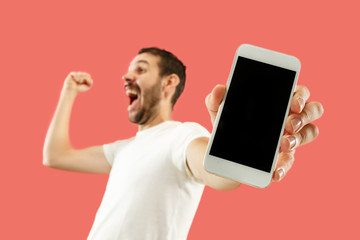 Young handsome man showing smartphone screen over coral background with a surprise face. Human...