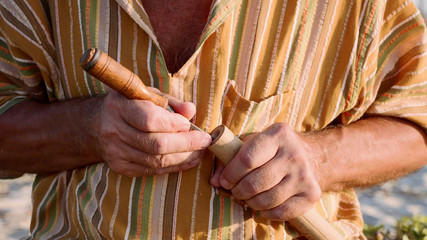 Senior man sits on the beach and making flute by hands, close-up. © alexeg84