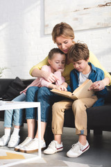 smiling mother with cute little kids sitting on couch and reading book