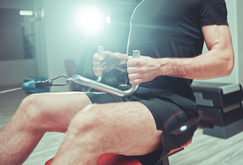 Man is training with equipment rowing training machine  by pulling handles while sitting. Exercising back muscles with weight at gym.