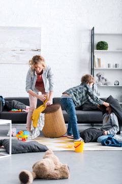 Tired Mother Putting Toys In Basket While Naughty Kids Fighting With Pillows