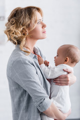 side view of thoughtful mother holding adorable infant child and looking away at home