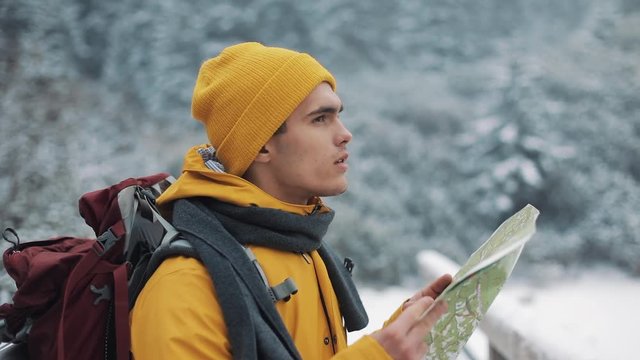 Young attractive man wearing yellow winter clothing holding map standing on the bridge and checking the route . Beautiful mountains in winter time in the background