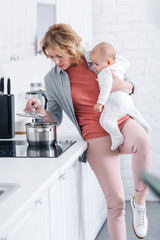 exhausted mother holding adorable infant child and cooking in kitchen