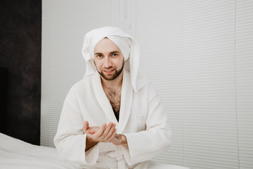 Relaxed bearded young man in a towel on his head and white coat with moisturizer on the face. Photo of smiling man on beige background. Grooming himself after shower