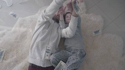 Joyful barded father and happy son lying at the white fur carpet catching money falling from above. Entertainment dad and child. Father-child relationship. Slow motion.