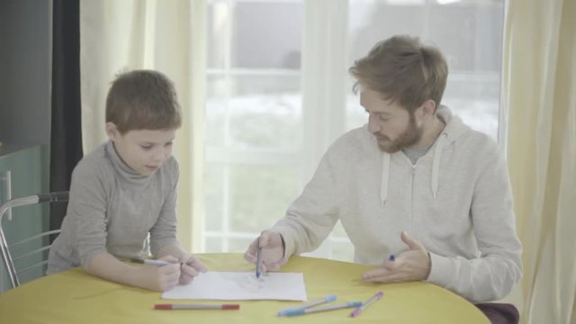 Bearded smilling father and his little son paint on paper sitting in living room at the table. Father-child relationship.