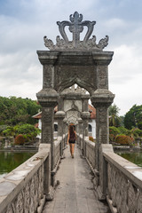 Blond girl walking away on a stone bridge in historical Bali hindu temple, surrounded by water