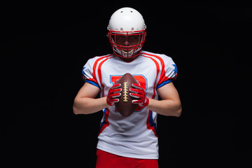 American football player wearing helmet holding ball in front of him on black background