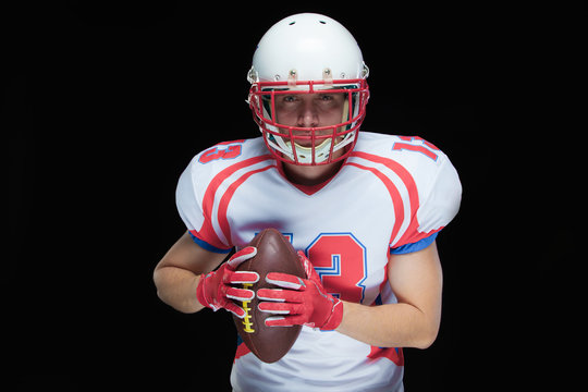 American football player wearing helmet posing with ball on black background