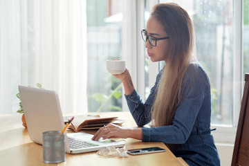 Attractive young Asian woman drinking coffee and working on laptop