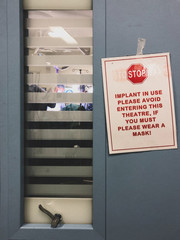 Sign outside operating theatre doors - do not enter - with surgeons visible  (blurred) through window blinds. 