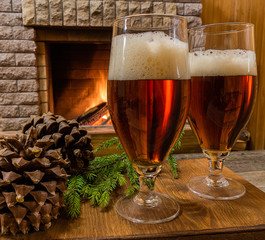 Two beer glasses, cones, christmas tree branch against cozy firepace.