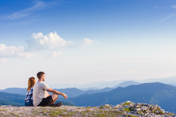 Naklejka na ściany i meble Sitting on the top of the world. A couple enjoying the view on a peak of the mountain having a zen moment, cliff at sunset. Success, winners, zen, tranquility, meditation.