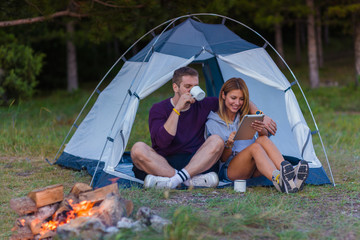 Young couple drinking coffee, enjoying the mountain view and checking the tablet with bonfire in the sunset