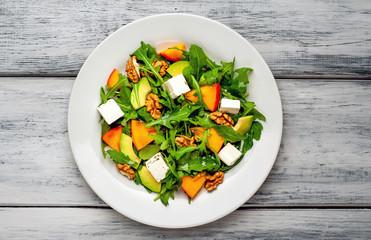 Arugula salad, persimmon, walnuts, avocados, feta cheese on a white plate on a wooden table