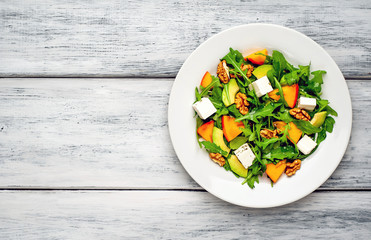 Arugula salad, persimmon, walnuts, avocados, feta cheese on a white plate on a wooden table
