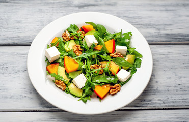 Arugula salad, persimmon, walnuts, avocados, feta cheese on a white plate on a wooden table