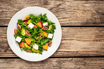 Arugula salad, persimmon, walnuts, avocados, feta cheese on a white plate on a wooden table