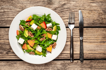 Arugula salad, persimmon, walnuts, avocados, feta cheese on a white plate on a wooden table