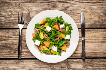 Arugula salad, persimmon, walnuts, avocados, feta cheese on a white plate on a wooden table