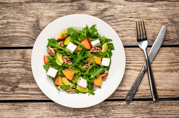 Arugula salad, persimmon, walnuts, avocados, feta cheese on a white plate on a wooden table