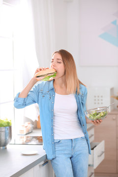 Beautiful young woman eating sandwich instead of salad in kitchen. Failed diet
