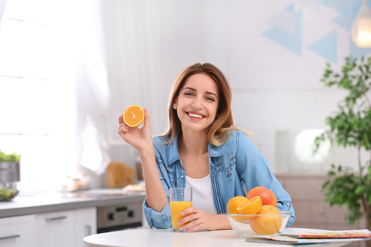 Happy young woman with glass of juice and orange at table in kitchen. Healthy diet