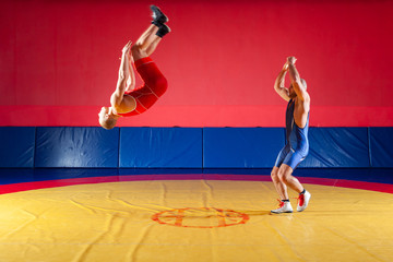 Two strong wrestlers in blue and red tights make flips and jumps on the yellow wrestling track in the gym. A young man makes a grab.
