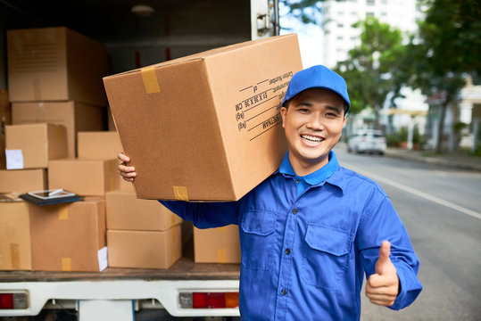 Happy young delivery man with big cardboard box showing thumbs-up and smiling at camera