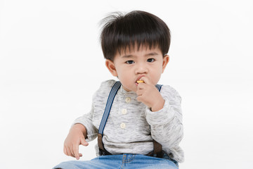 Studio portrait of cute, adorable, Asian toddler boy wearing denim overalls, long sleeve T-shirt, orange shoes, sitting spread legs on the floor, enjoy eating snack, on isolated white background